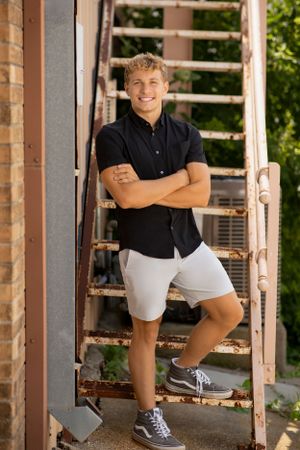 A person in a black shirt and white shorts poses confidently on rustic metal stairs outdoors on a sunny day.