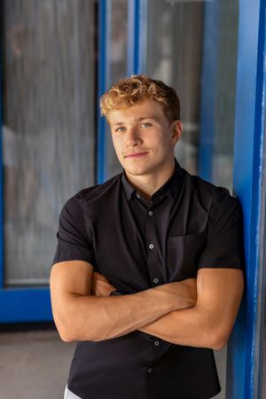 Young athletic person in black button-down shirt poses confidently with crossed arms against blue door frame.