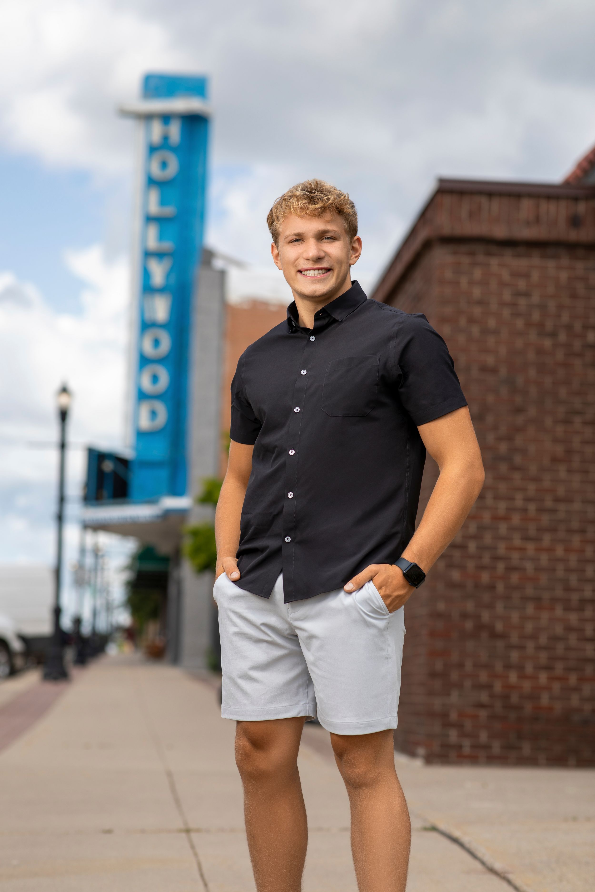 A person in a black shirt and grey shorts poses casually on a sidewalk with a Hollywood sign visible in the background.
