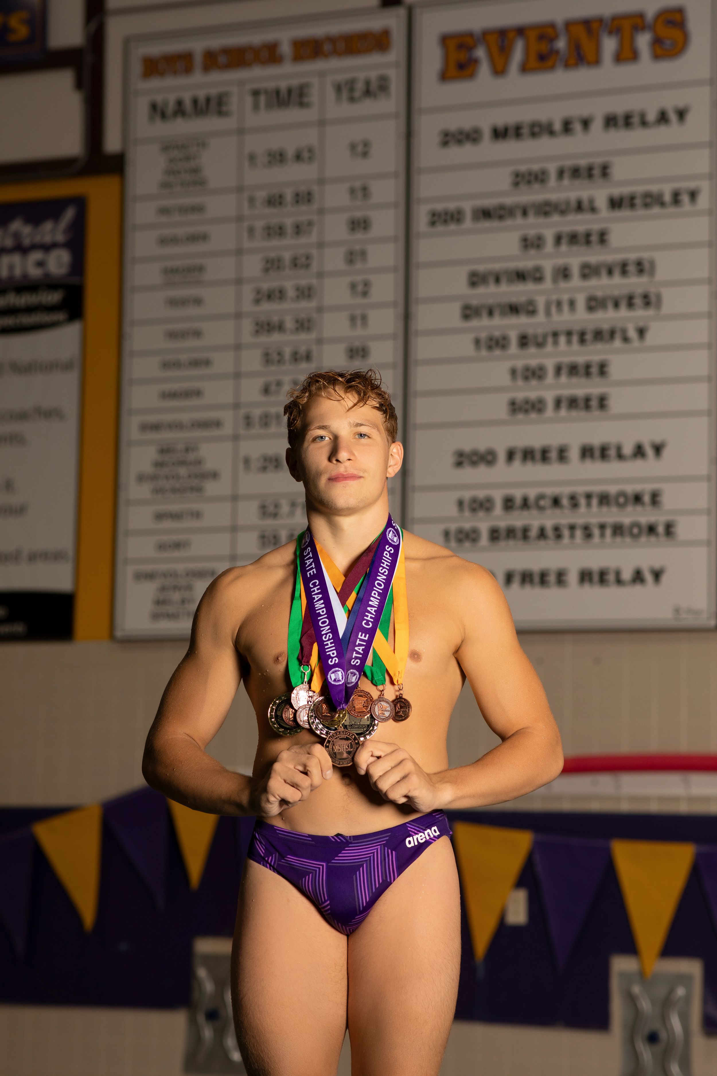 Athlete stands proudly displaying multiple swimming medals in front of school record board at pool.
