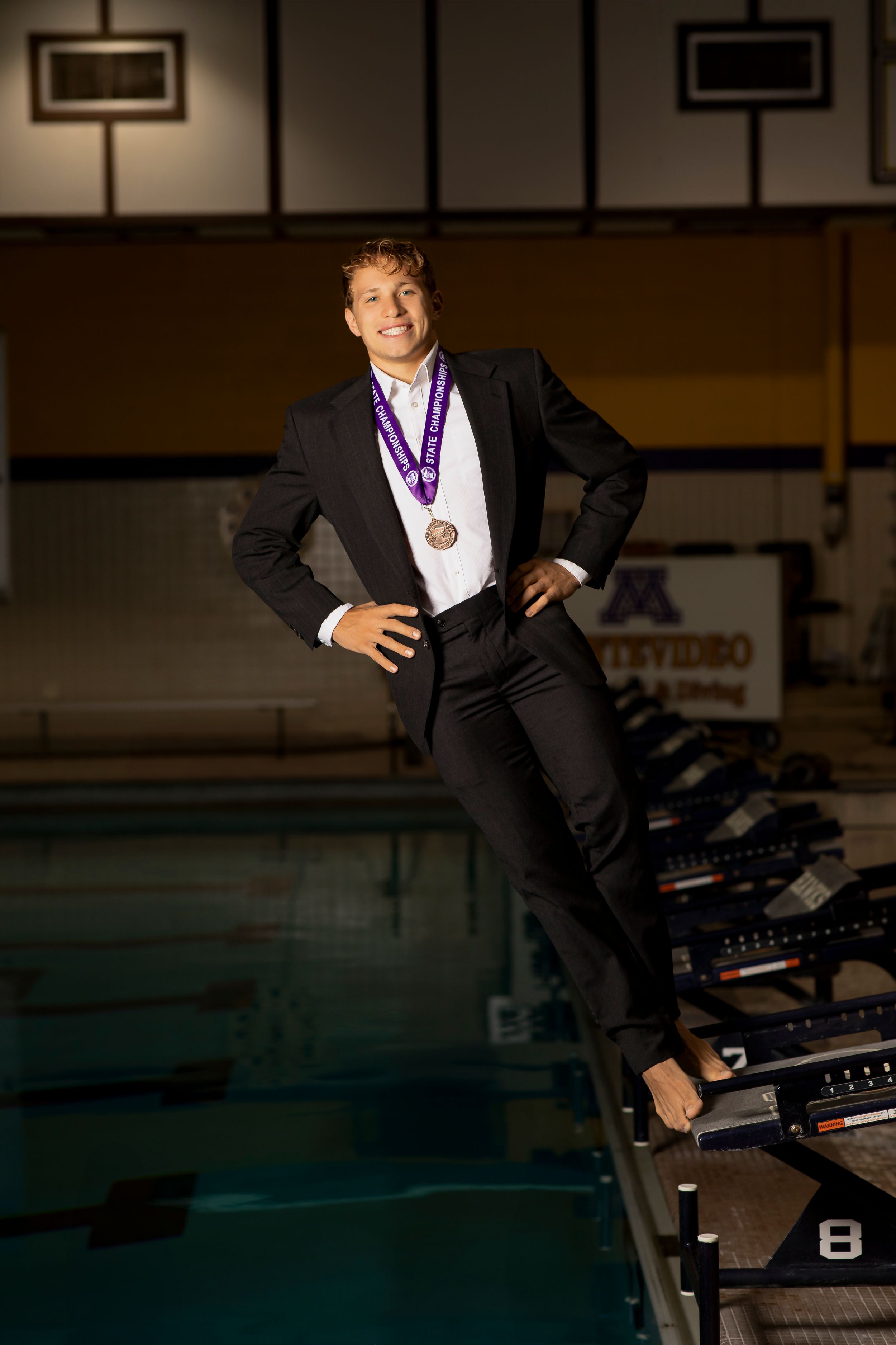 Athlete in formal suit poses with medal while standing on starting blocks at indoor swimming pool.