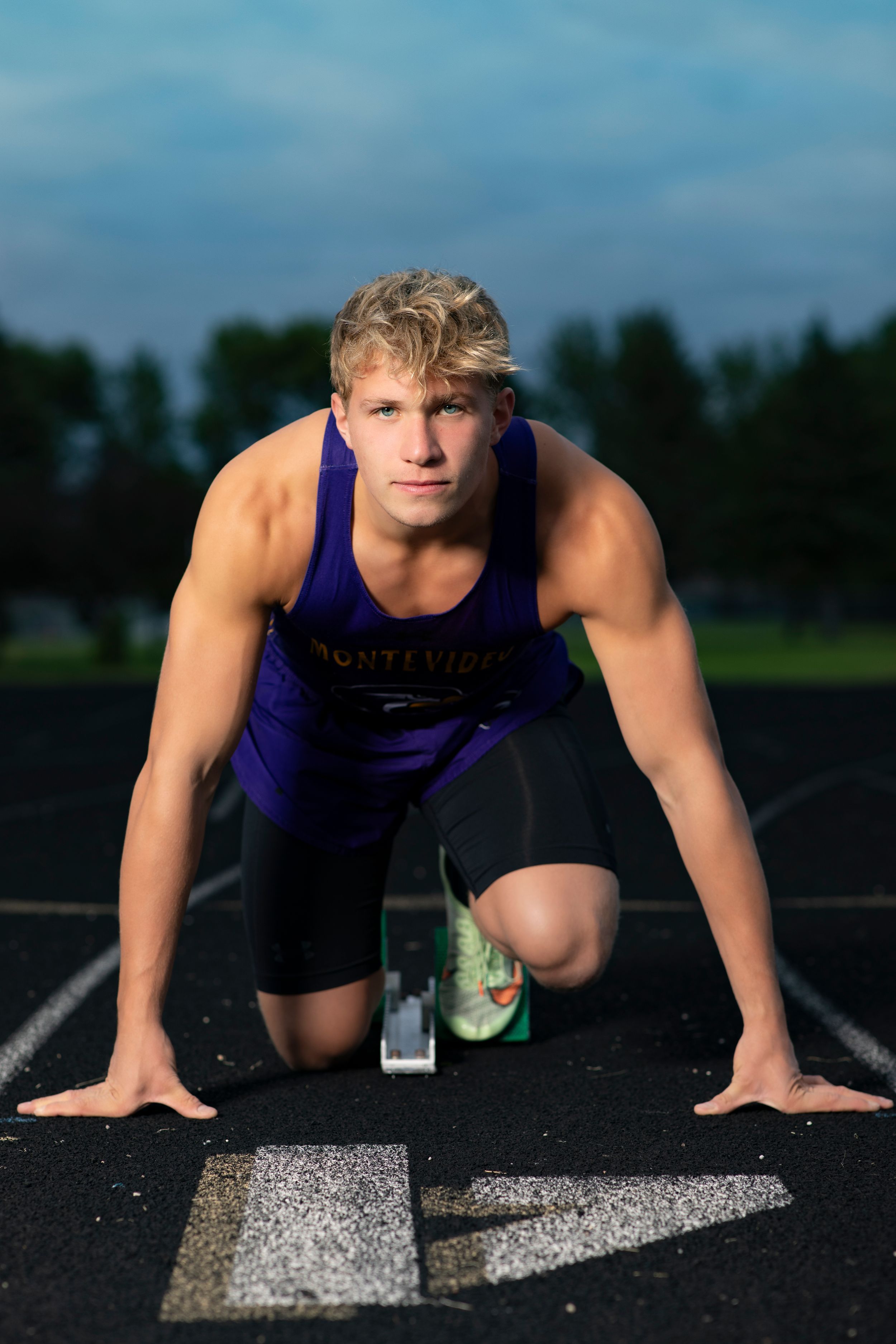 Athletic runner in purple tank top and black shorts at starting position on outdoor track during dusk.