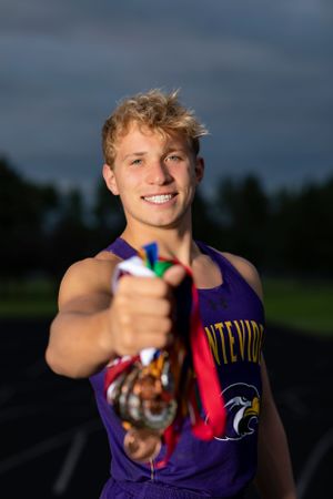 High school athlete in purple jersey holds up medal with proud smile during outdoor track portrait session.