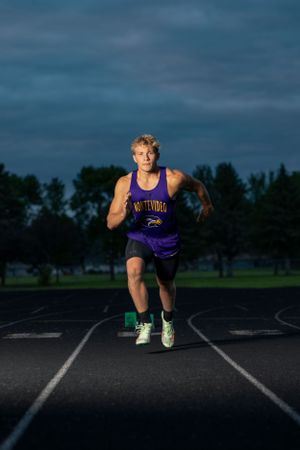 An athlete in a purple tank top sprints on a dark track during evening training.