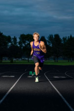 High school athlete in purple uniform sprints down track lane at dusk with trees in background.