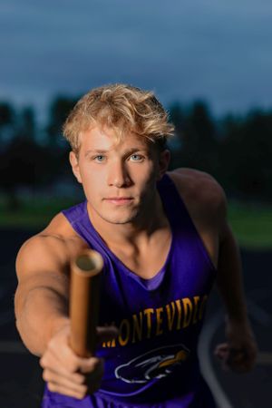 Track and field athlete wearing a purple Montevideo uniform extends a relay baton during a race at dusk.