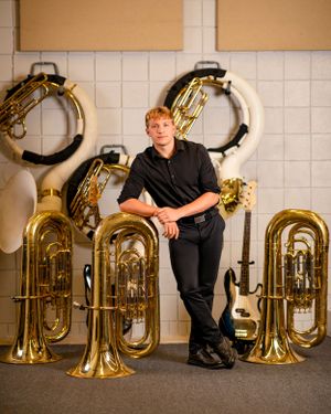 A person stands beside multiple shiny brass tubas and sousaphones displayed against a white tiled wall.