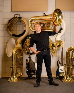 A musician stands surrounded by brass instruments including tubas, electric guitars and drums in a rehearsal room.