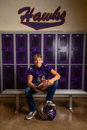 High school football player in purple Hawks jersey sits on locker room bench with football and helmet beside him.