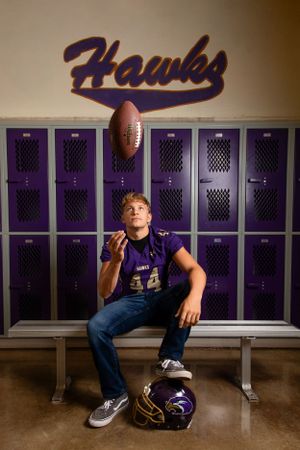 A football player sits in front of purple lockers tossing a football while wearing a Hawks jersey and jeans.