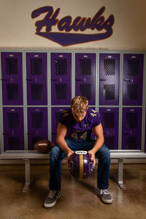 Football player sits on locker room bench with purple lockers and Hawks logo on wall in background.