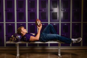 Student lying on locker room bench holding football while wearing jeans and athletic shoes in purple-lit environment.
