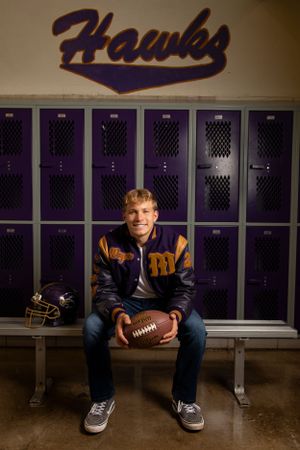 High school athlete in varsity jacket sits on bench holding football in front of purple lockers with Hawks team logo above.