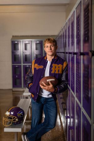 High school football athlete poses in varsity jacket holding football beside purple lockers and helmet.