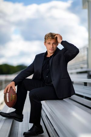 Professional portrait of a young athlete in dark suit sitting on stadium bleachers holding a football on a sunny day.