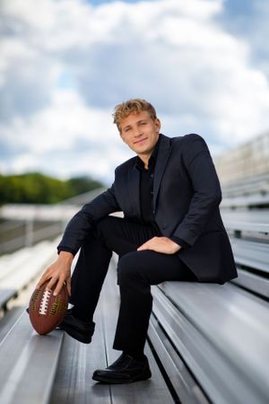 Athletic senior portrait on metal bleachers shows student in dark suit holding football against cloudy sky background.