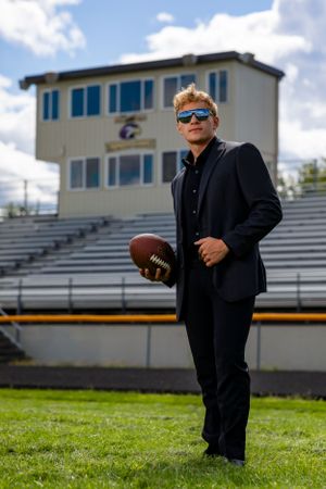 A well-dressed person in dark attire stands on a football field holding a football with stadium bleachers in the background.