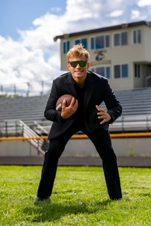 Athletic figure in sunglasses and black attire poses with football on grass field with bleachers in background.
