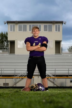 Football player in purple uniform stands with crossed arms on field in front of bleachers and press box.