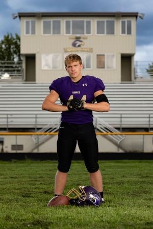 High school football player in purple jersey stands on field with helmet and ball by bleachers under blue sky.