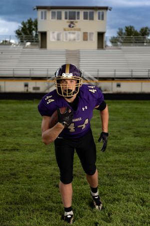 High school football player in purple uniform running with the ball on field with stadium bleachers in background.