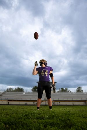 Football player in purple jersey number 44 tosses ball in air on grassy field under dramatic cloudy sky.