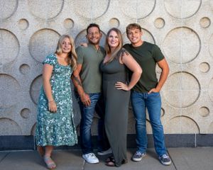 A group of four people pose together against a gray textured wall with circular pattern design in an indoor setting.