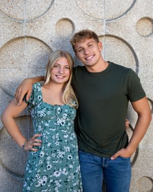A couple stands together against a decorative concrete wall, with the woman wearing a floral dress and the man in a dark shirt.