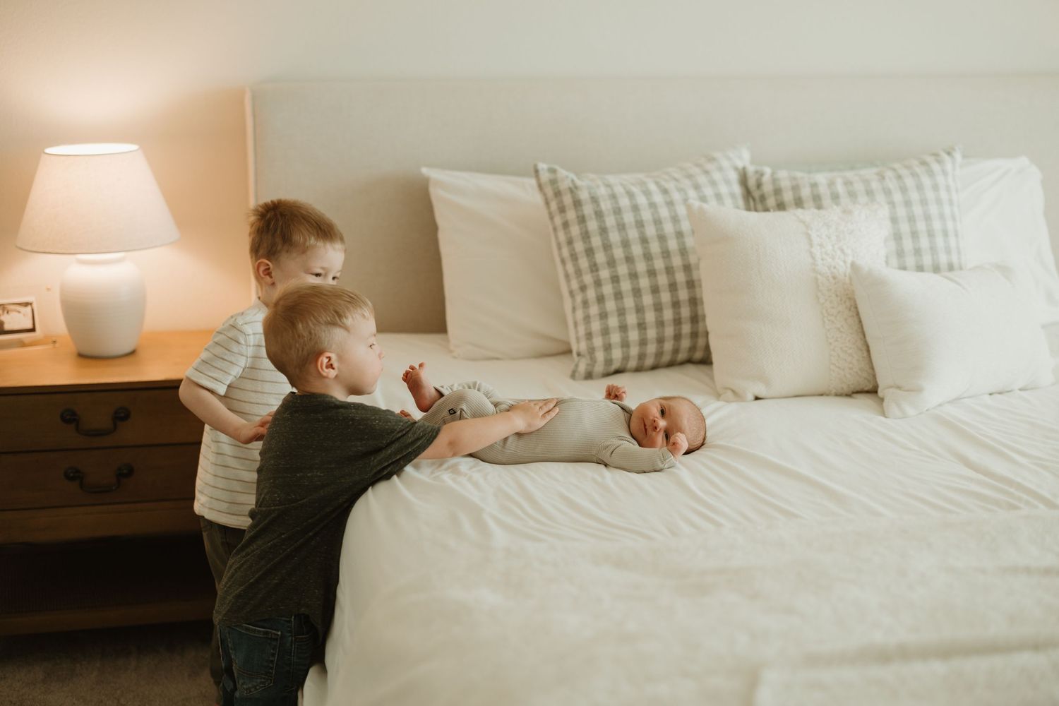 Two young children playing together on a white bed with gingham pillows and a wooden nightstand with table lamp.