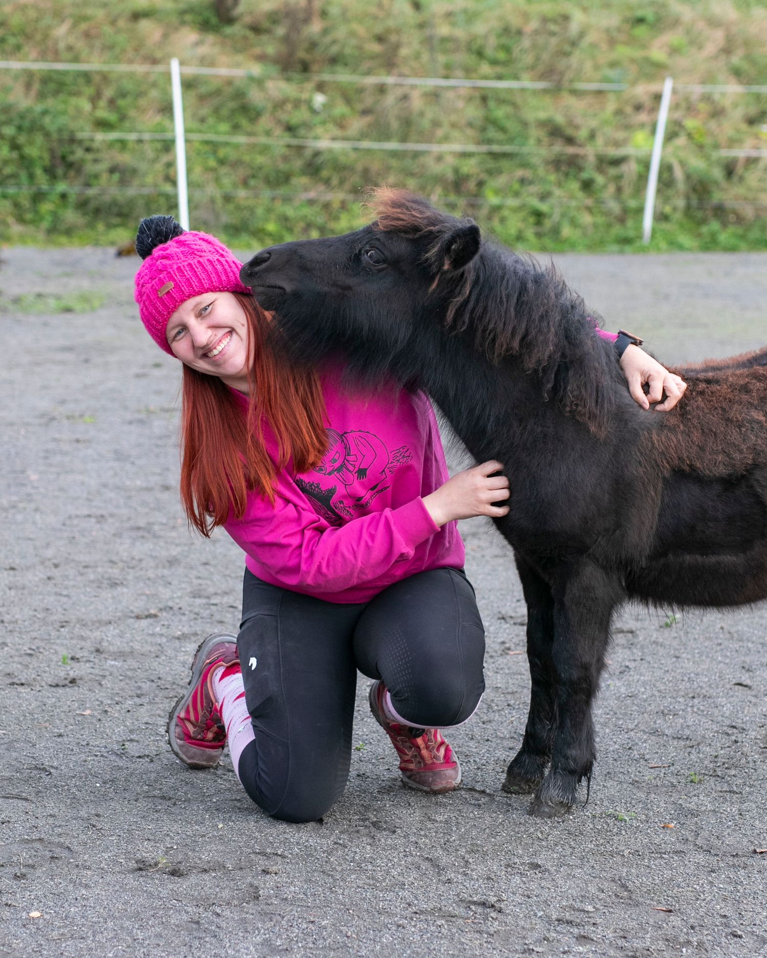 A bright pink-clad figure kneels next to a small black pony in an outdoor paddock area.
