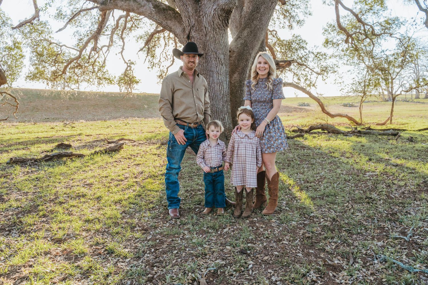 Family of four posing under a large oak tree in a green field, dressed in country-style outfits for a portrait.