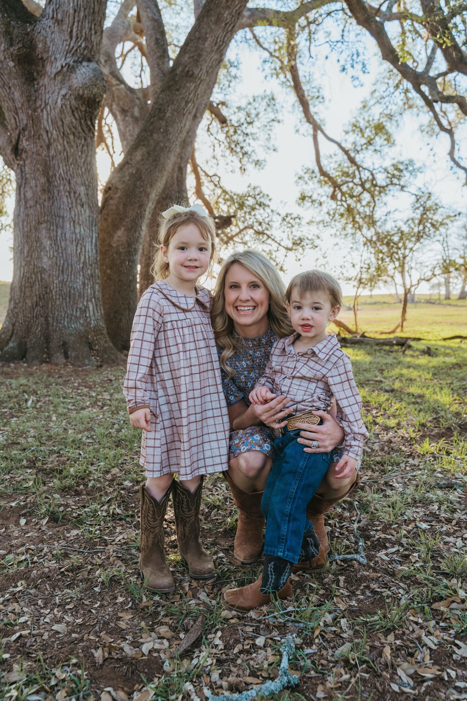 A smiling blonde woman kneels outdoors holding a toddler boy beside a young girl in plaid dress and cowboy boots near a large oak tree.