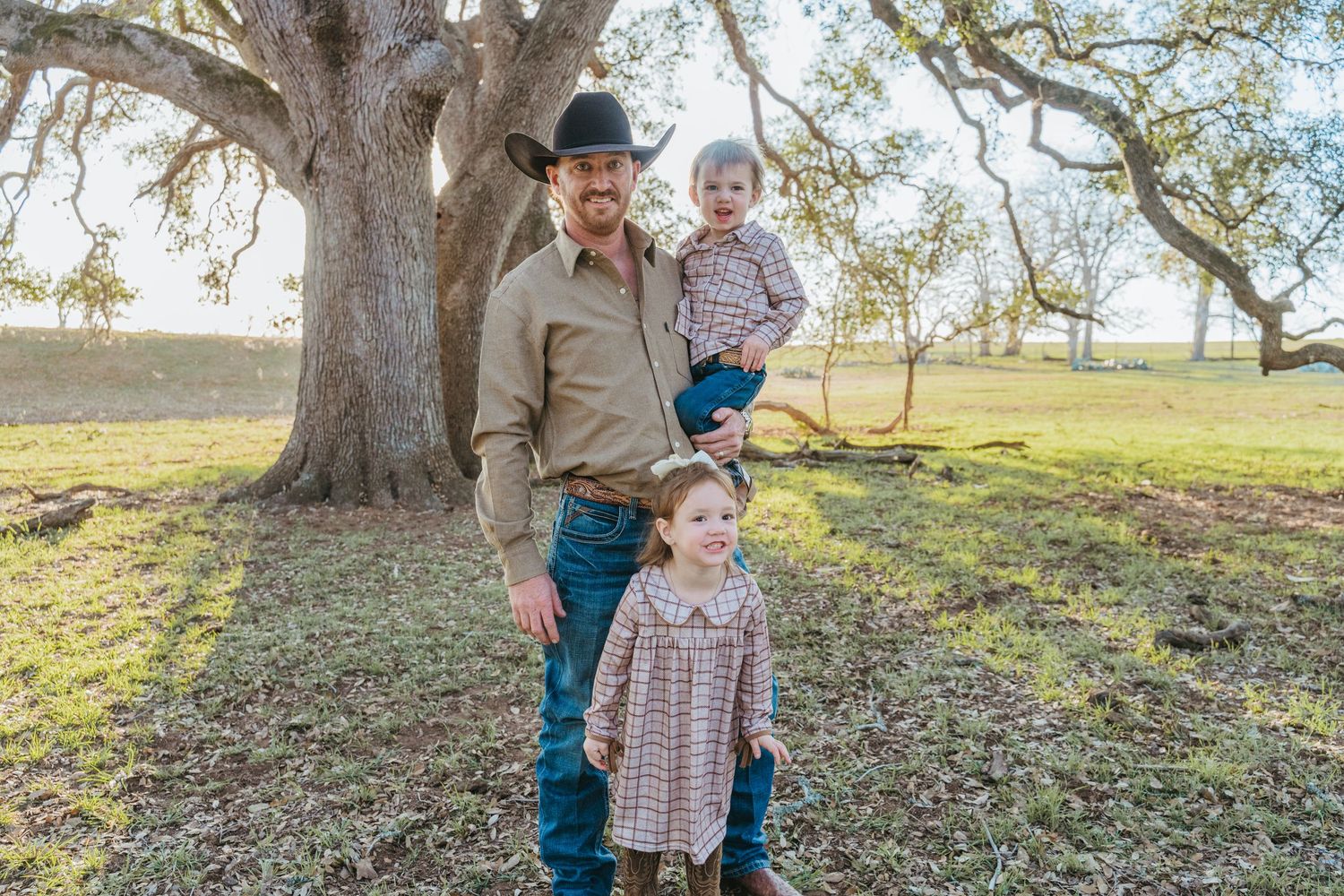 A father in a cowboy hat holds his toddler son while his young daughter stands beside him near a large oak tree.