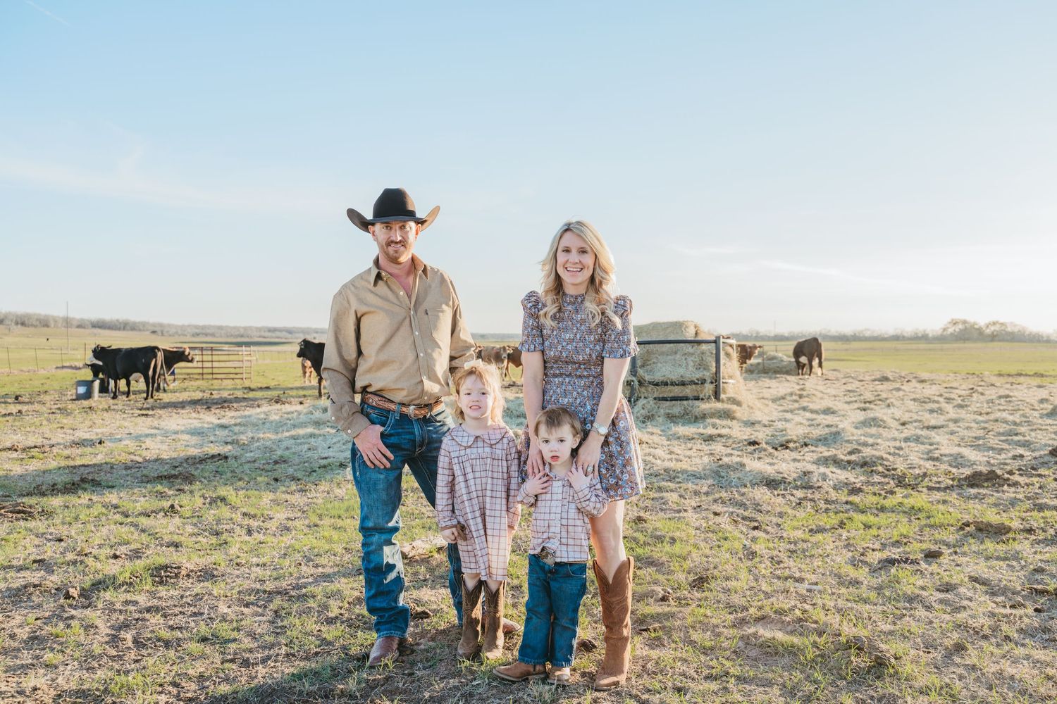 Family of four posing on a ranch with cattle in background, father wearing cowboy hat and boots on sunny day.