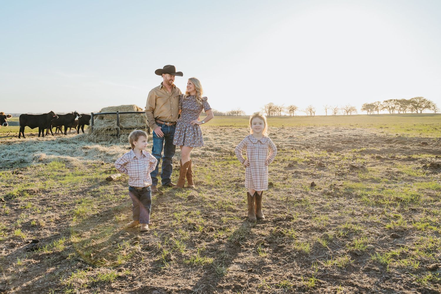 Family of four stands in a sunny cattle ranch field, father in cowboy hat with wife and two young children.