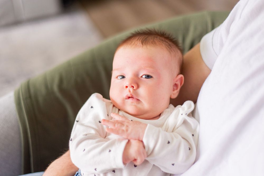 A baby in a white patterned onesie rests on a cozy green couch.