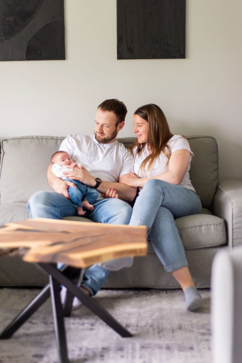 A couple sits together with their newborn baby on a grey couch in a modern living room with black wall art.