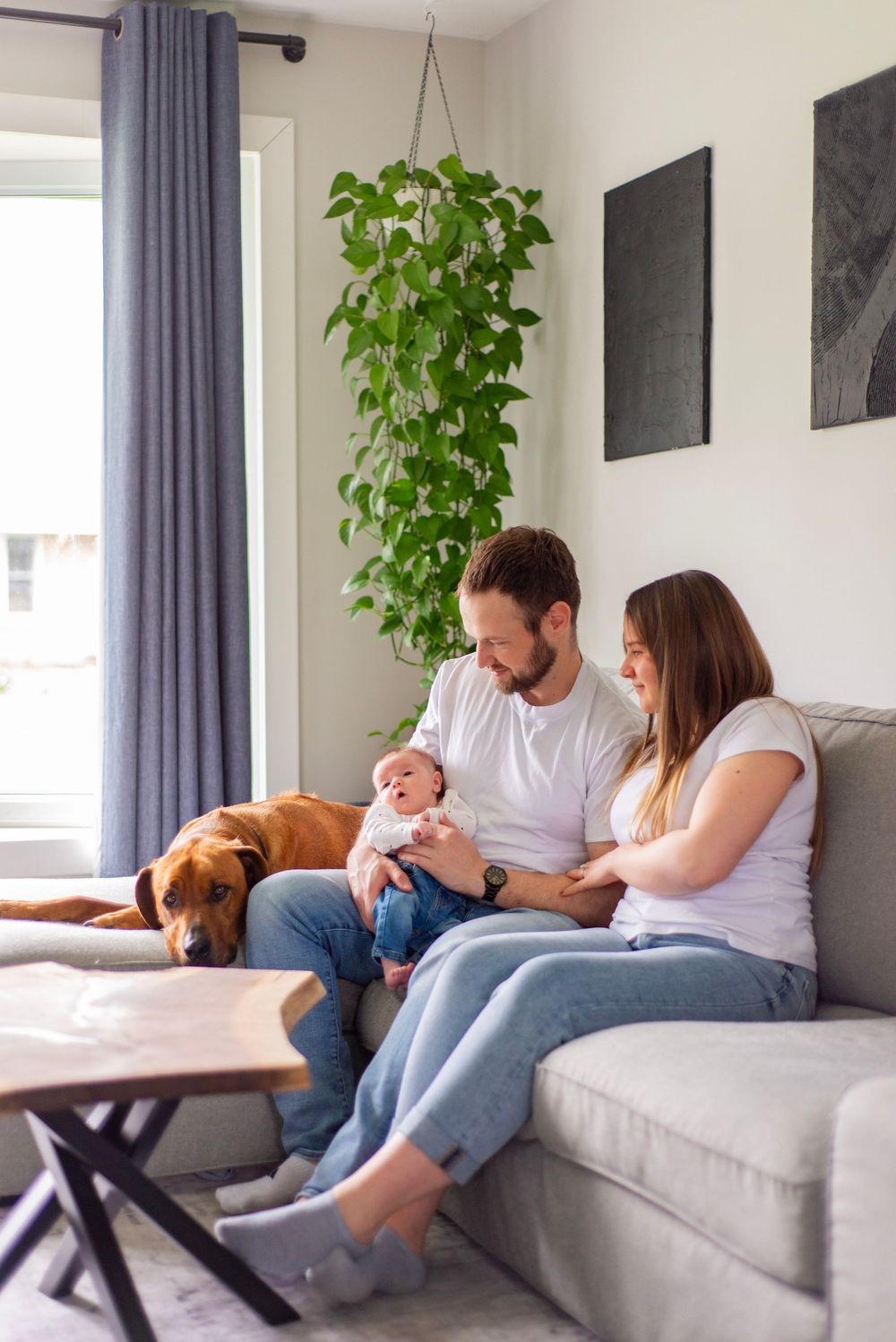 A young family relaxes on a grey couch in their living room with a brown dog and hanging potted plant nearby.