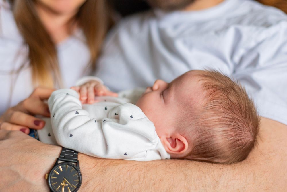 Sleeping baby wearing white sleepwear rests comfortably on adult's arm with a wristwatch visible.