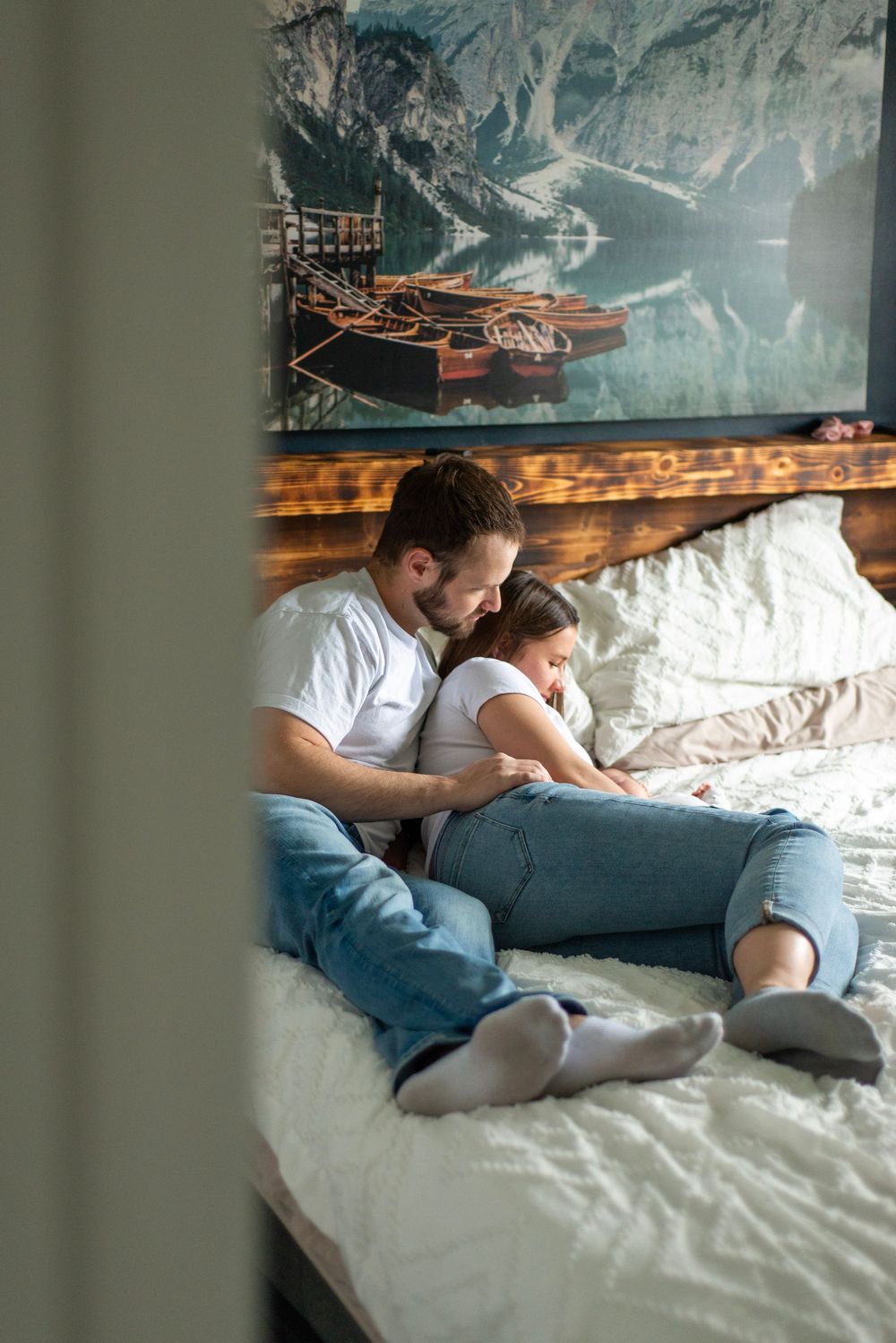 Two people relax together on a cozy white bed with a scenic mountain lake artwork displayed on the wall above.