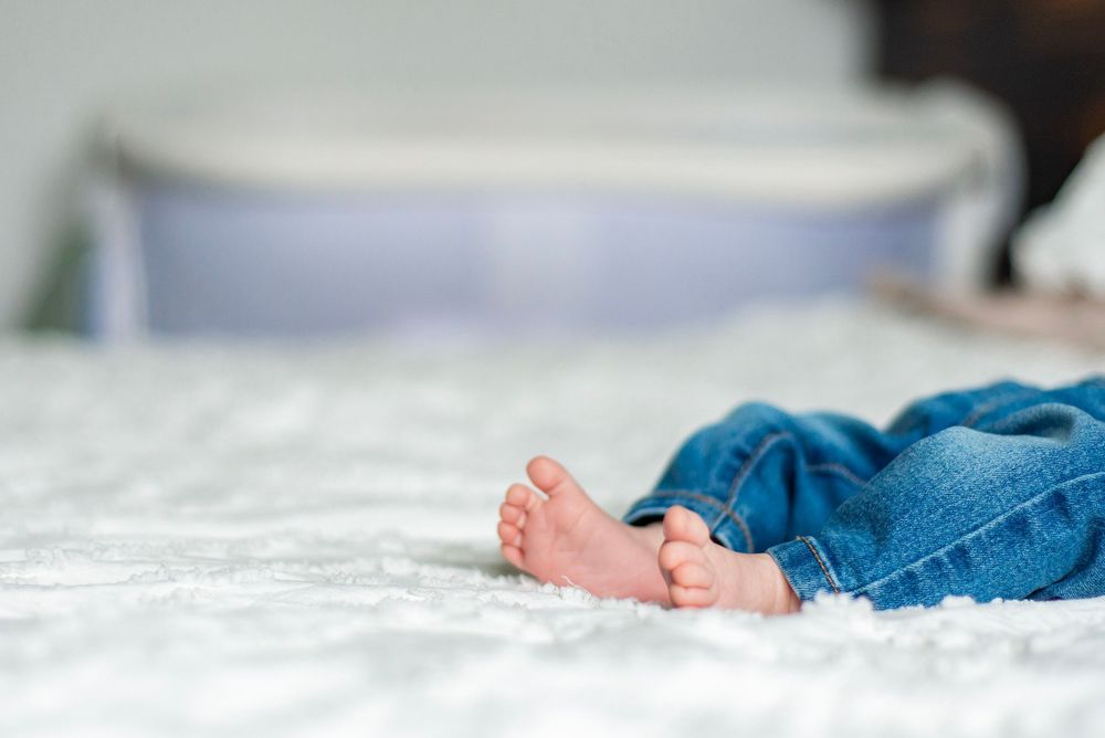 Small bare feet peek out from blue jeans while resting on white bedding in a soft, intimate photograph.