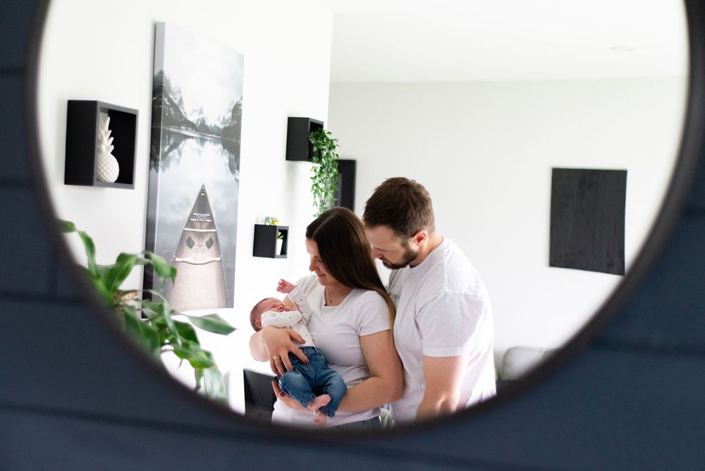 Framed mirror reflection shows parents holding their newborn baby in a minimalist white room with black artwork and plants.
