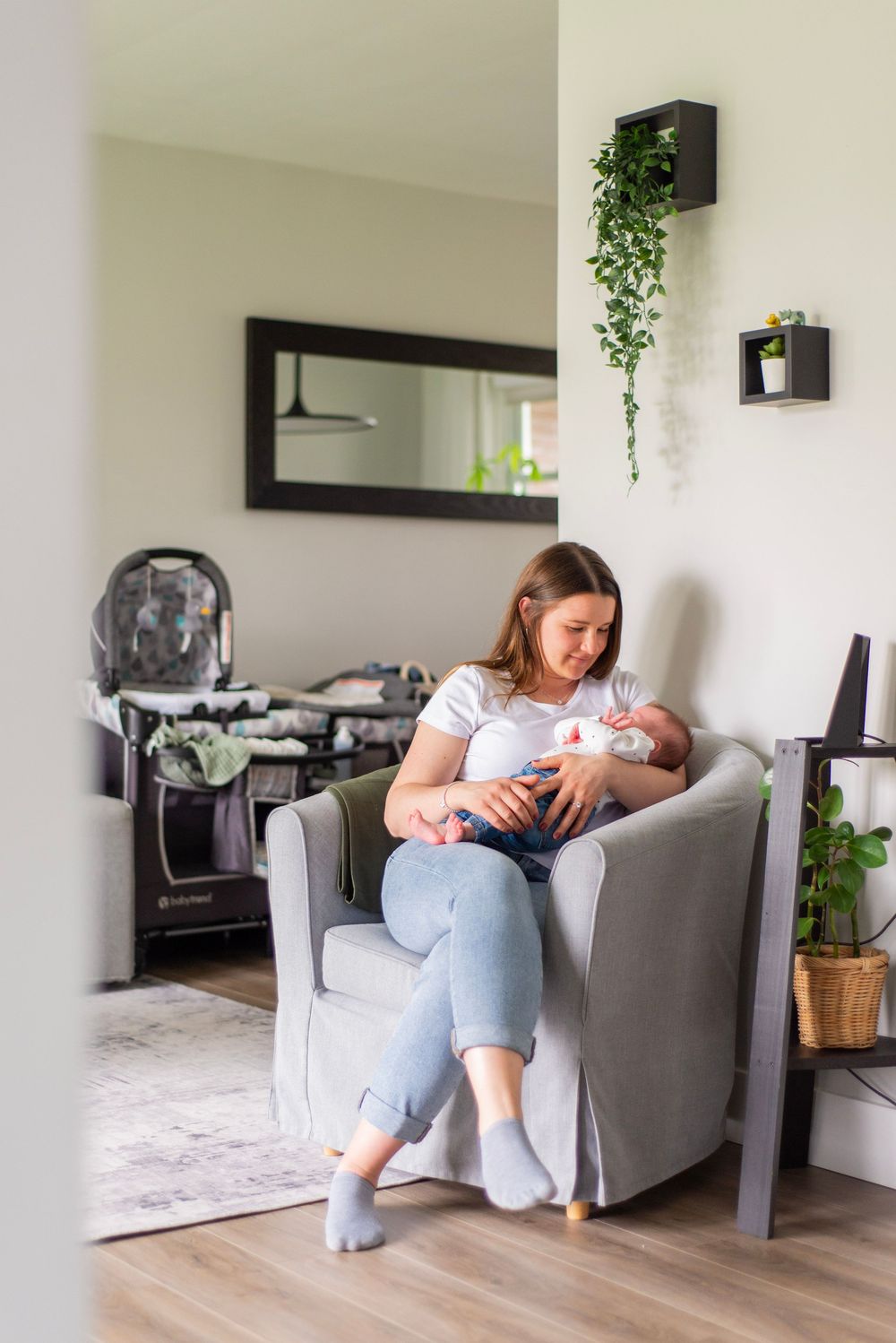Peaceful nursery corner with gray armchair, decorative mirror, and hanging plants creates a cozy feeding space.