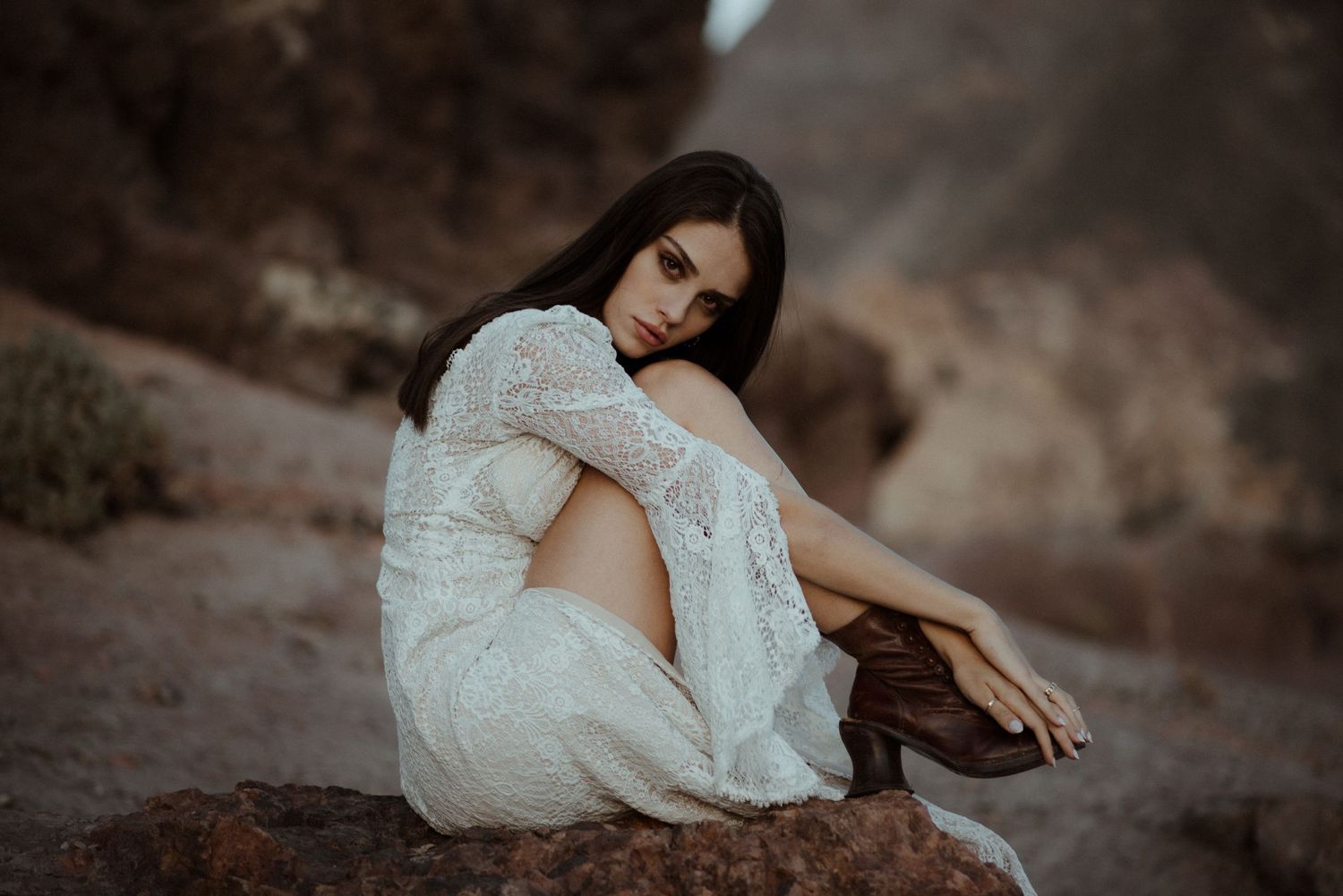 A person in white lace dress sits thoughtfully on desert rocks at dusk.