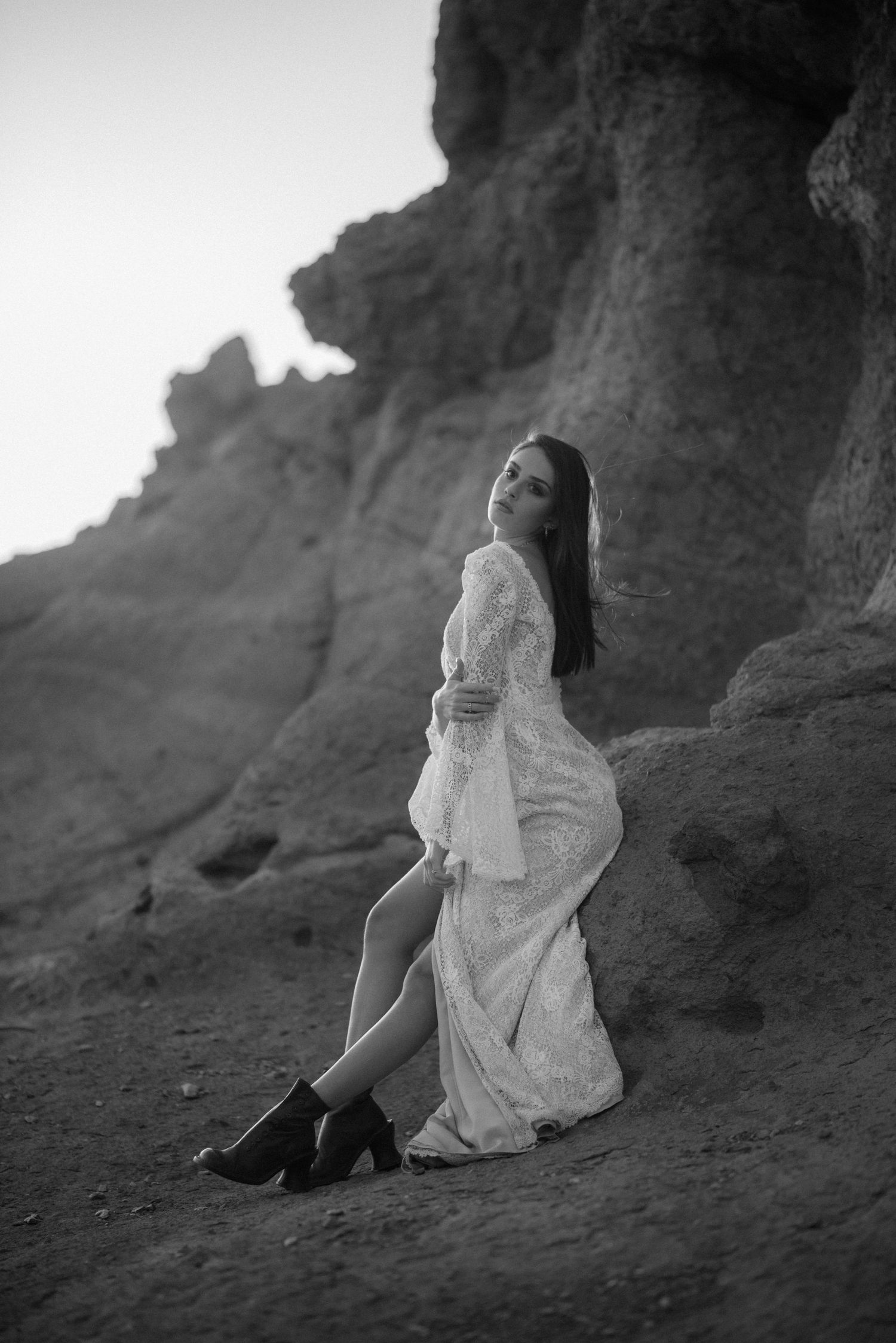 Black and white portrait of a bride in boots and lace dress against dramatic cliffs.