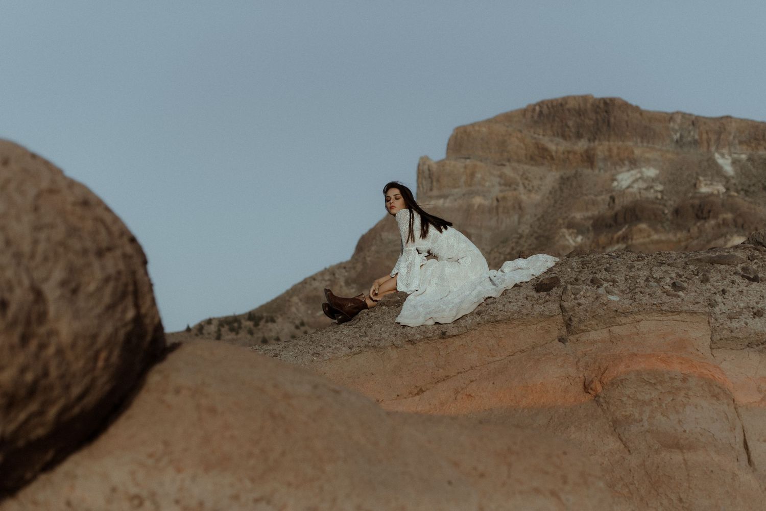 A white lace wedding dress flows elegantly against dramatic red rock formations at sunset.