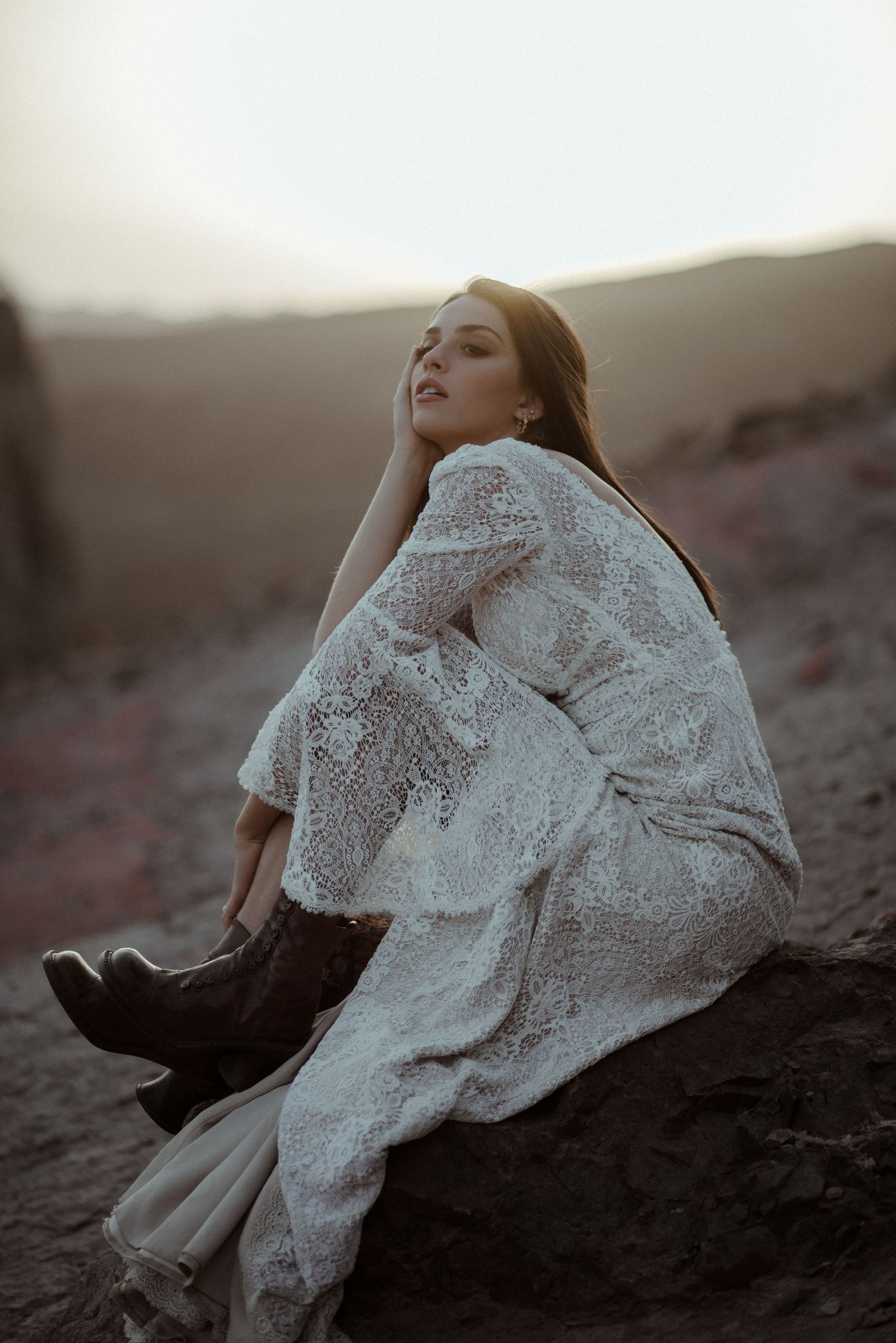 A dreamy outdoor portrait of someone in a flowing white lace dress and boots sitting on rocks.