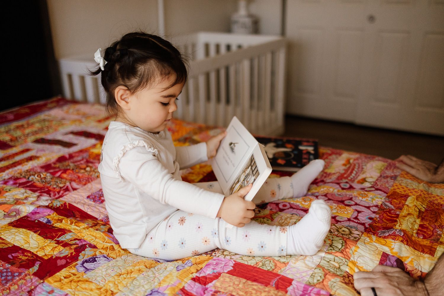 Young girl reads book on colorful bedspread at newborn photo session in San Jose