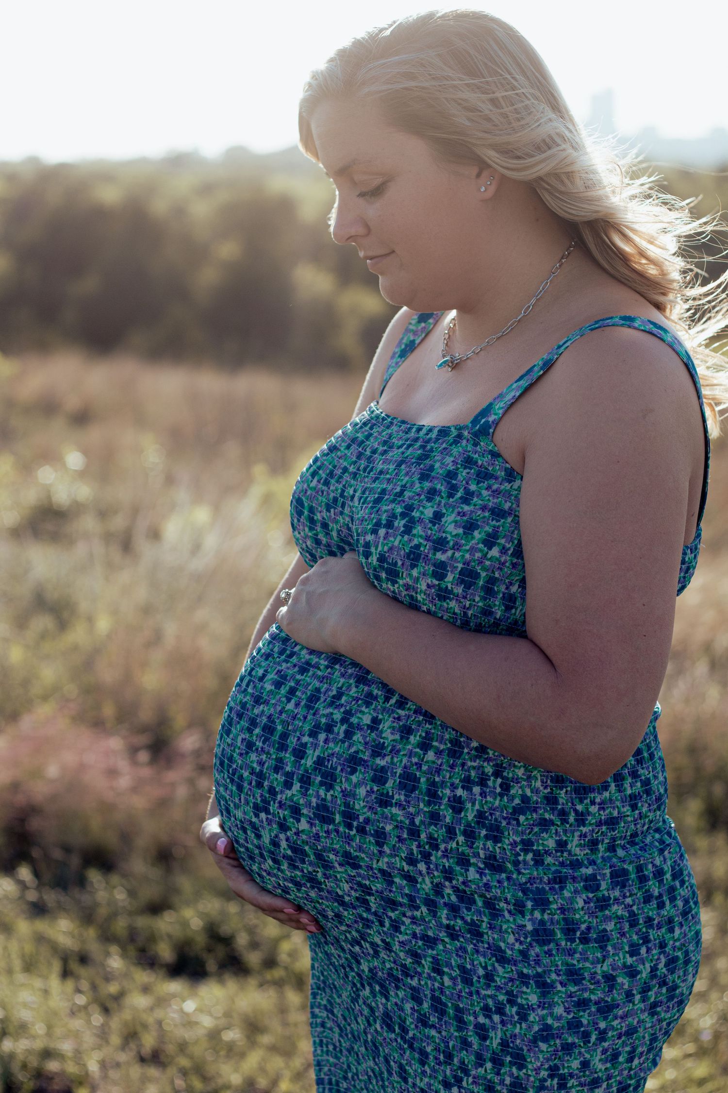 Side view of pregnant woman in blue floral dress caressing her baby bump while standing in golden sunlit field at sunset.