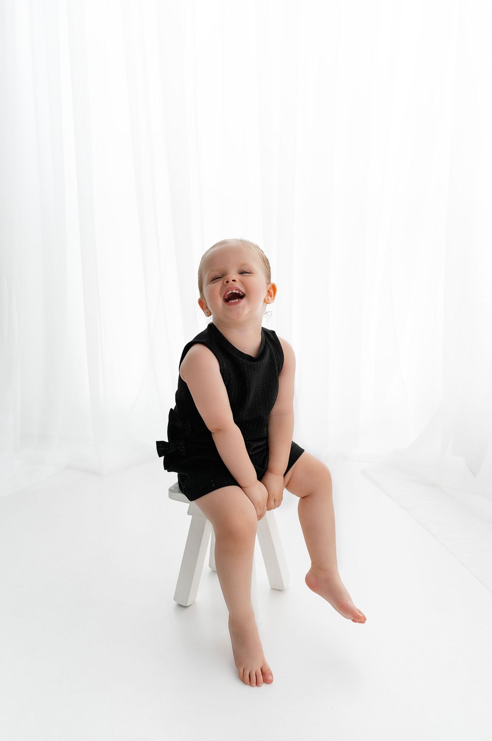 A joyful little one in a black outfit sits on a white stool against a bright white background.